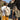 Rear view of a man and woman walking down a narrow stone street, both wearing large roll-top backpacks. One beige and one olive green.