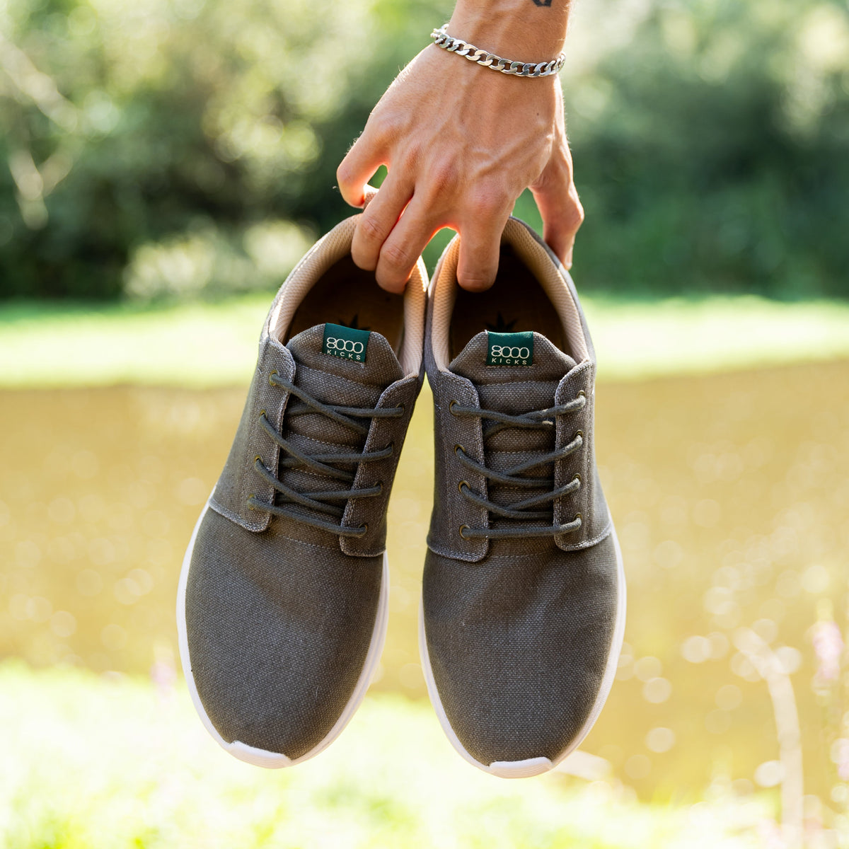 A tattooed hand holding a pair of explorer v2's against a blurred outdoor background of green foliage and water.