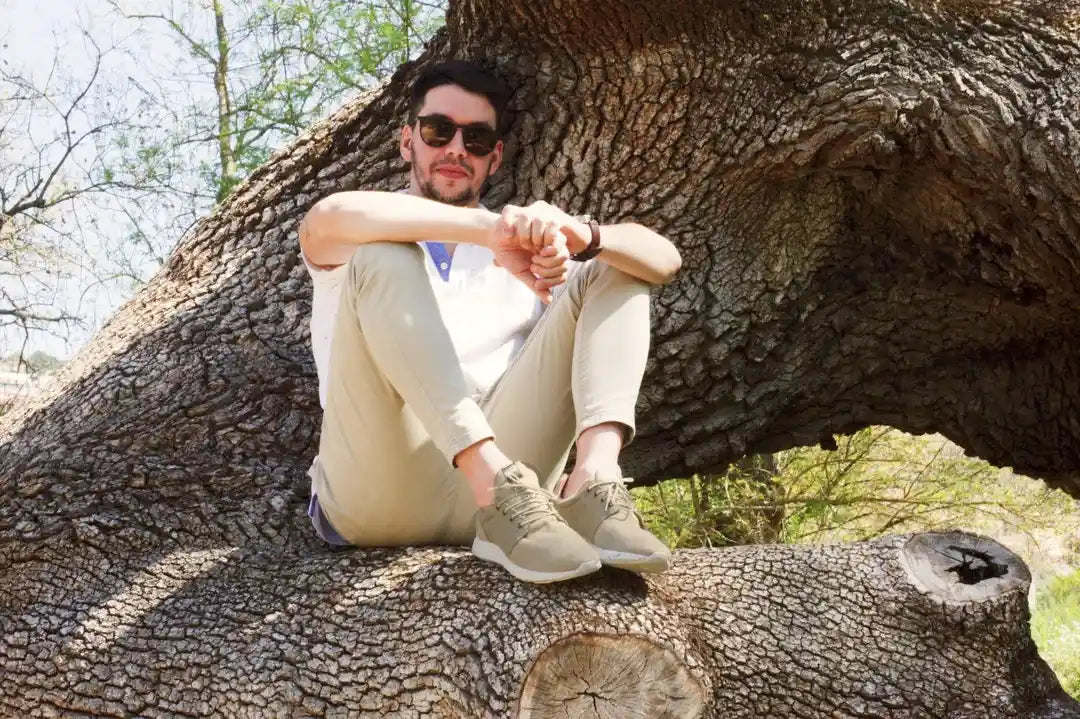 A man in sunglasses sits in an oak tree and shows off his beige and green hemp shoes.